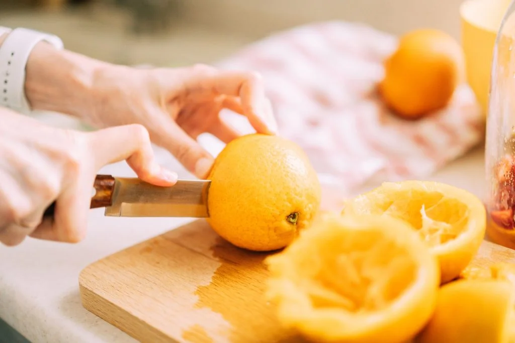 Female expertly slicing a fresh orange in half on a wooden cutting board, demonstrating proper technique for preparing nutrient-rich citrus fruits.